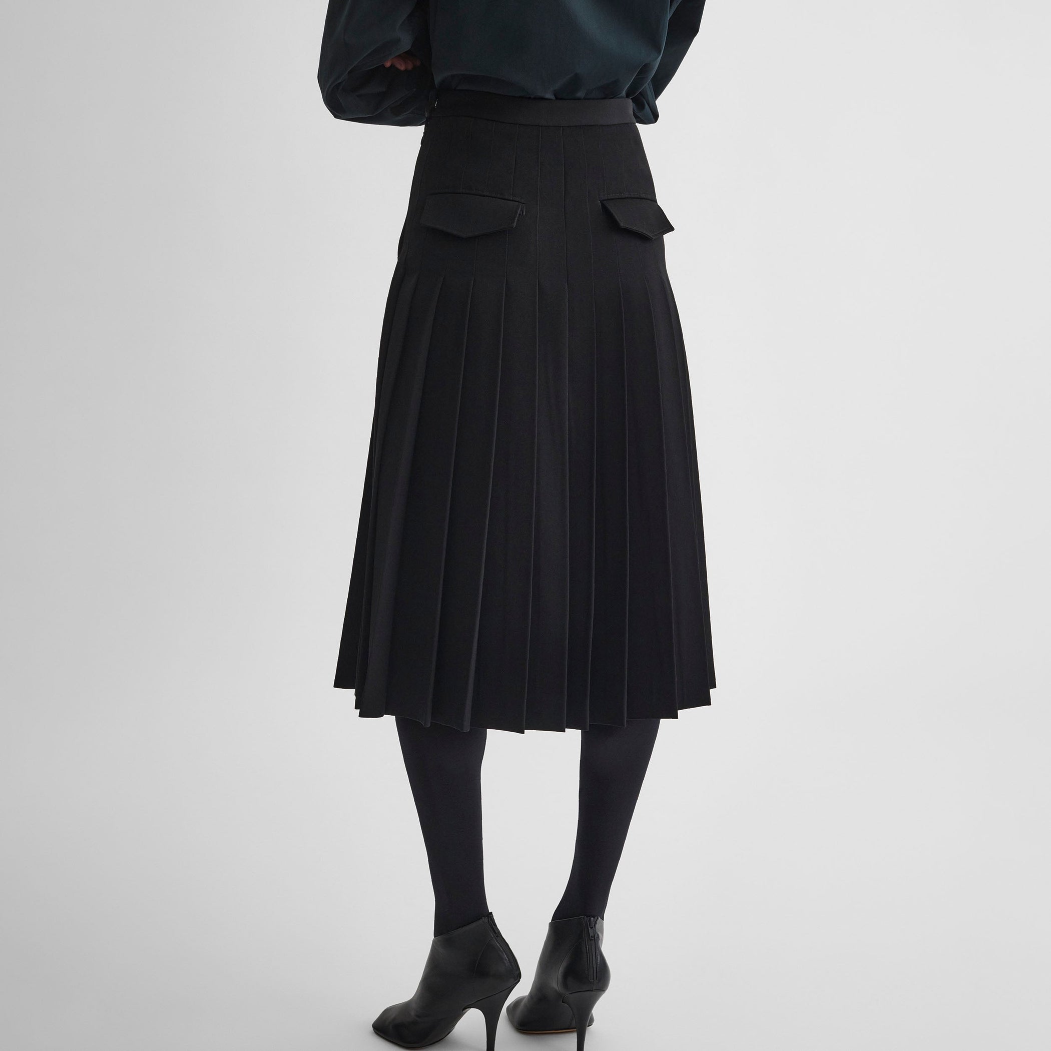 Back view of model wearing UNIFORM PLEATED SKIRT in black wool blend